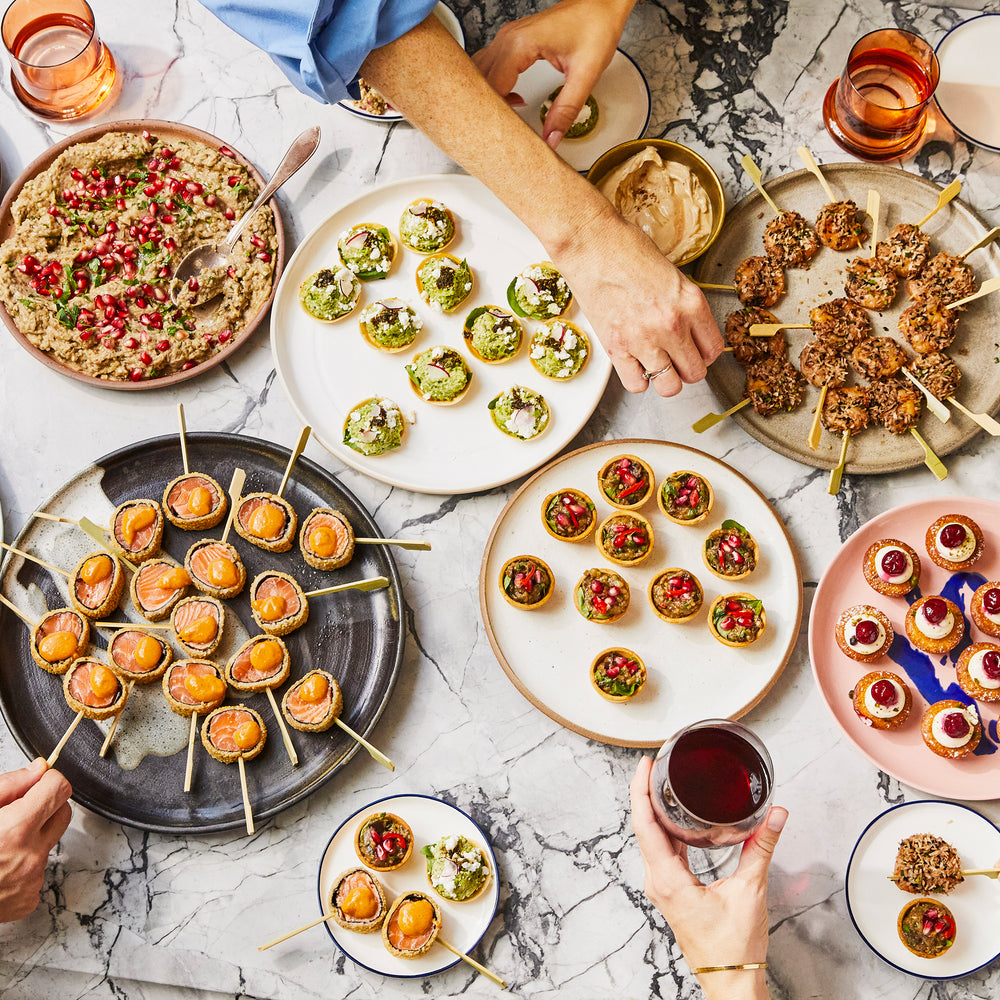 A table top set with canapes: seared salmon, sharp aubergine tart, pea and feta tart, prawn skewers and Persian love cake; burnt aubergine dip. Hands of people picking up the food. 