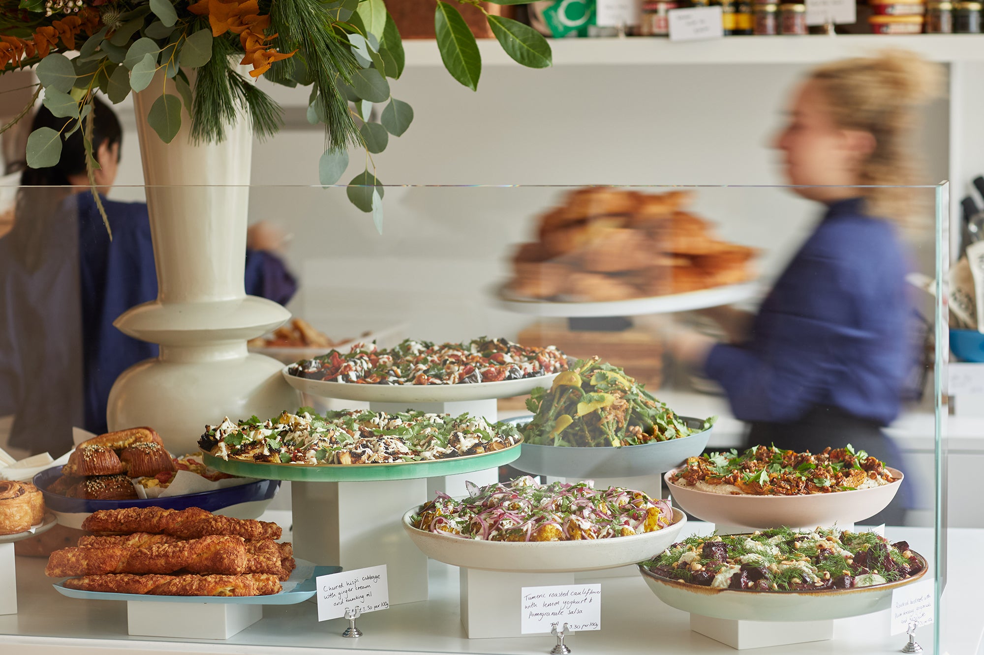 Display with salad platters, pastries and vase of flowers; servers begind.