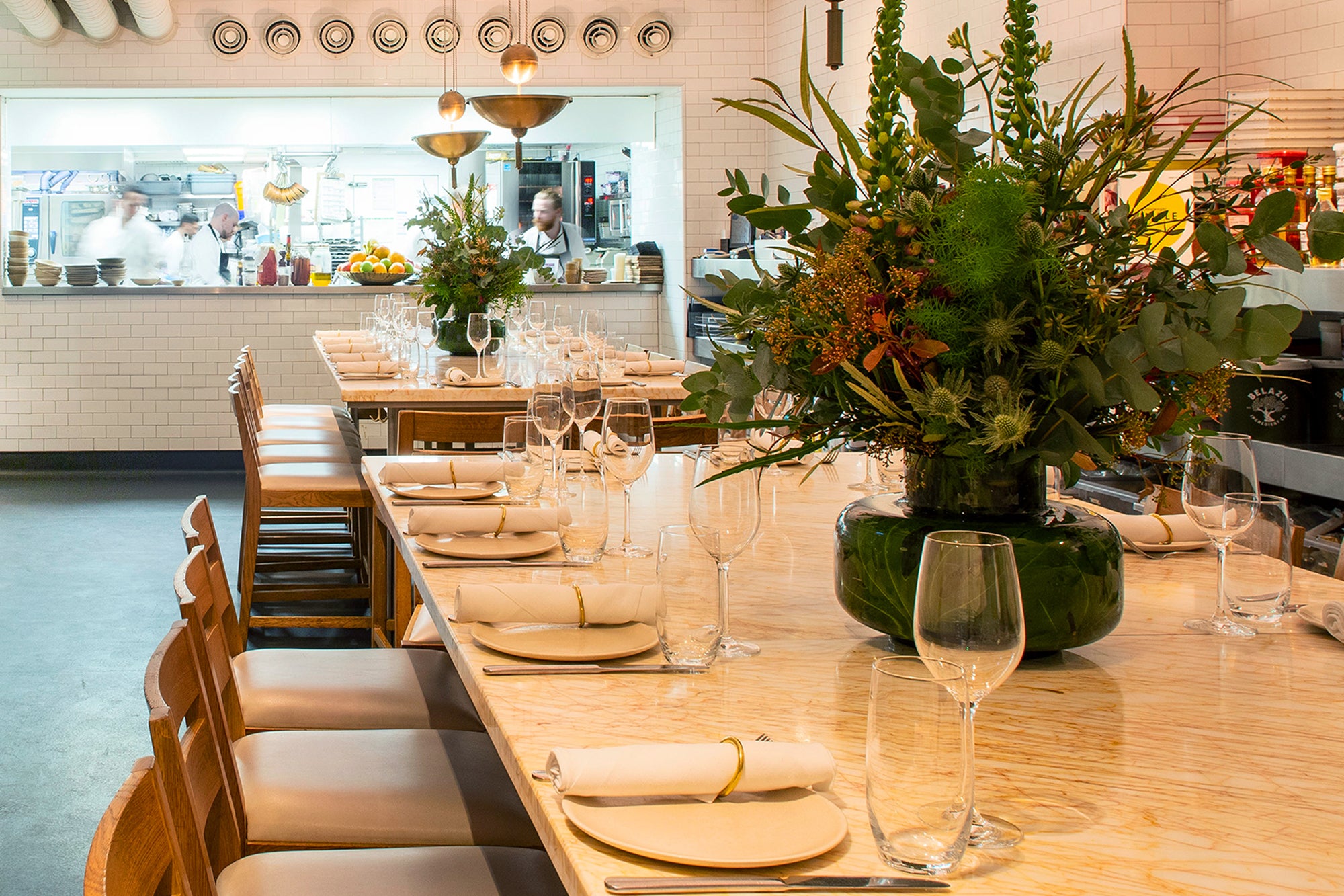 Two sharing table set up for dinner service and big flower vases. Open-plan kitchen at the background