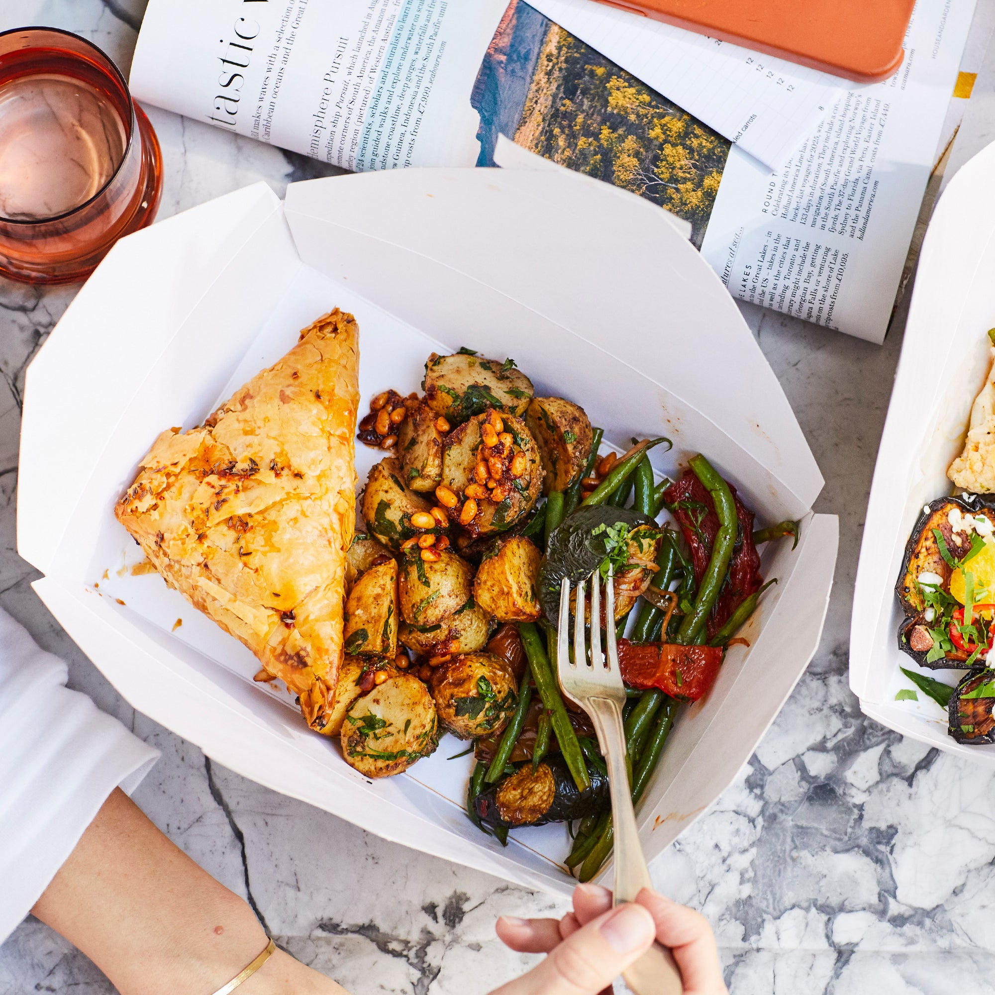 A box with with samosa, potato and green bean salads. A person holding a fork. A magazine on the table.